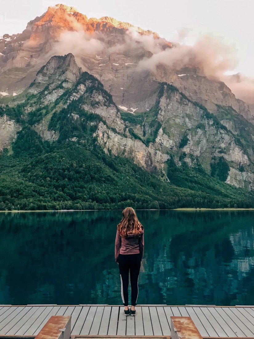 A woman with long hair stands with her back to the camera, standing on a small pier at a crystal blue lake. The lake extends out towards a small forest of green trees at the base of a towering Italian Alps mountain range.