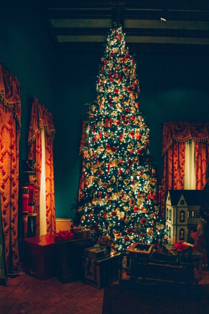 A giant Christmas tree is filled with lights and ornaments in a dark room. Boxes of wrapped Christmas presents surround the tree.