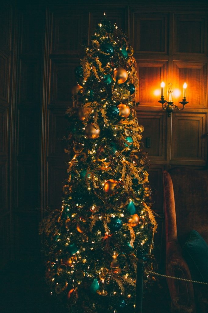 Another tall Christmas tree draped in lights, ornaments, and ribbons in a dark room, with warm light coming from a sconce on the wall.