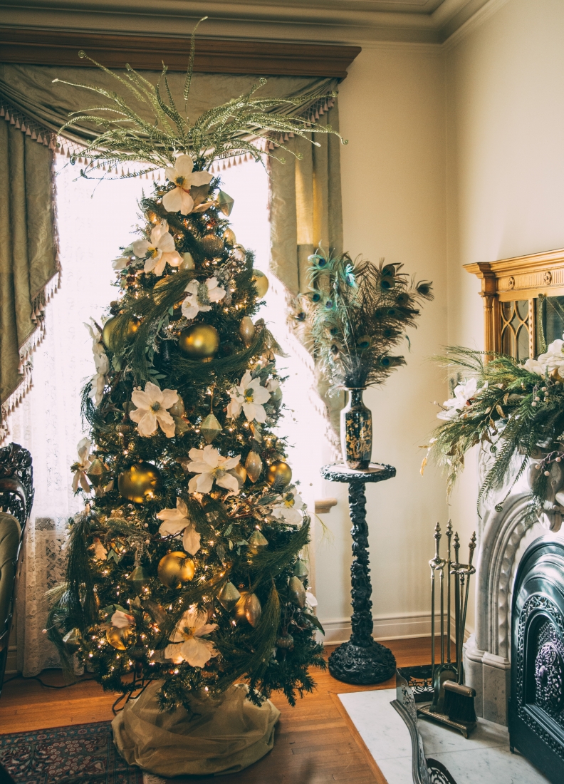 A large Christmas tree stands in front of a window beaming natural light into a bright room. The tree is decorated with white flowers, gold ornaments, and green garlands.
