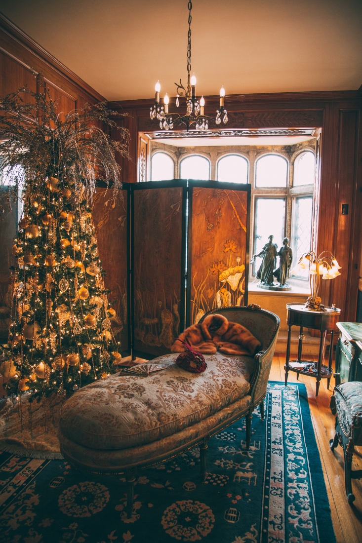 A dimly-lit sitting room with a vintage chaise lounge chair next to a decorated Christmas tree and a dressing screen. A large picture window in the back flows light into the warm room.