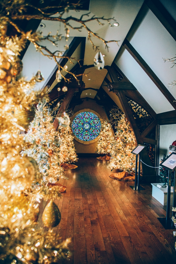 A long hallway with vaulted ceilings. The room is lined with brightly lit and decorated Christmas trees. At the end of the hallway is an ornate round stained glass window.