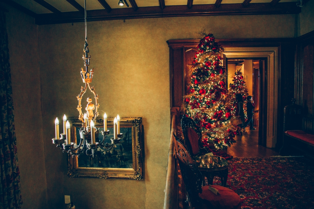 A chandelier hanging from a tall ceiling. Off to the side, a hallway is lined with decorated Christmas trees.