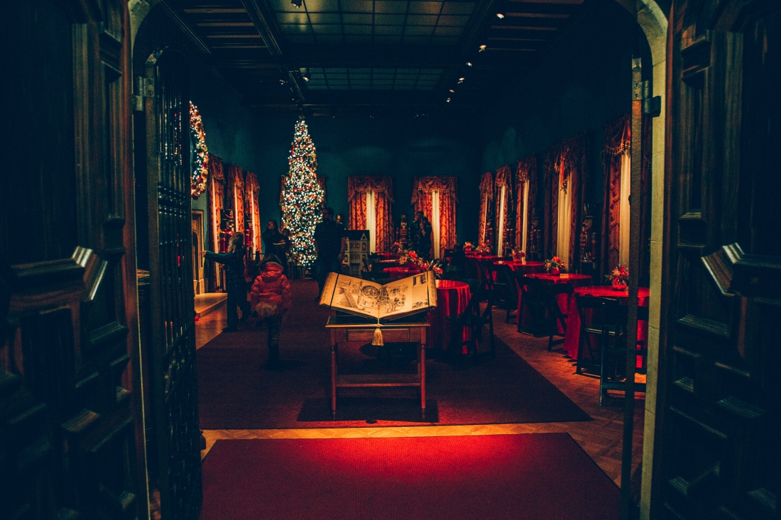 A look inside a dark, warmly lit room inside the Paine Art Center. A storybook is open on a table; behind it, a scene from the Nutcracker -- round tables set for dinner, and a large, bright Christmas tree in the back of the room.