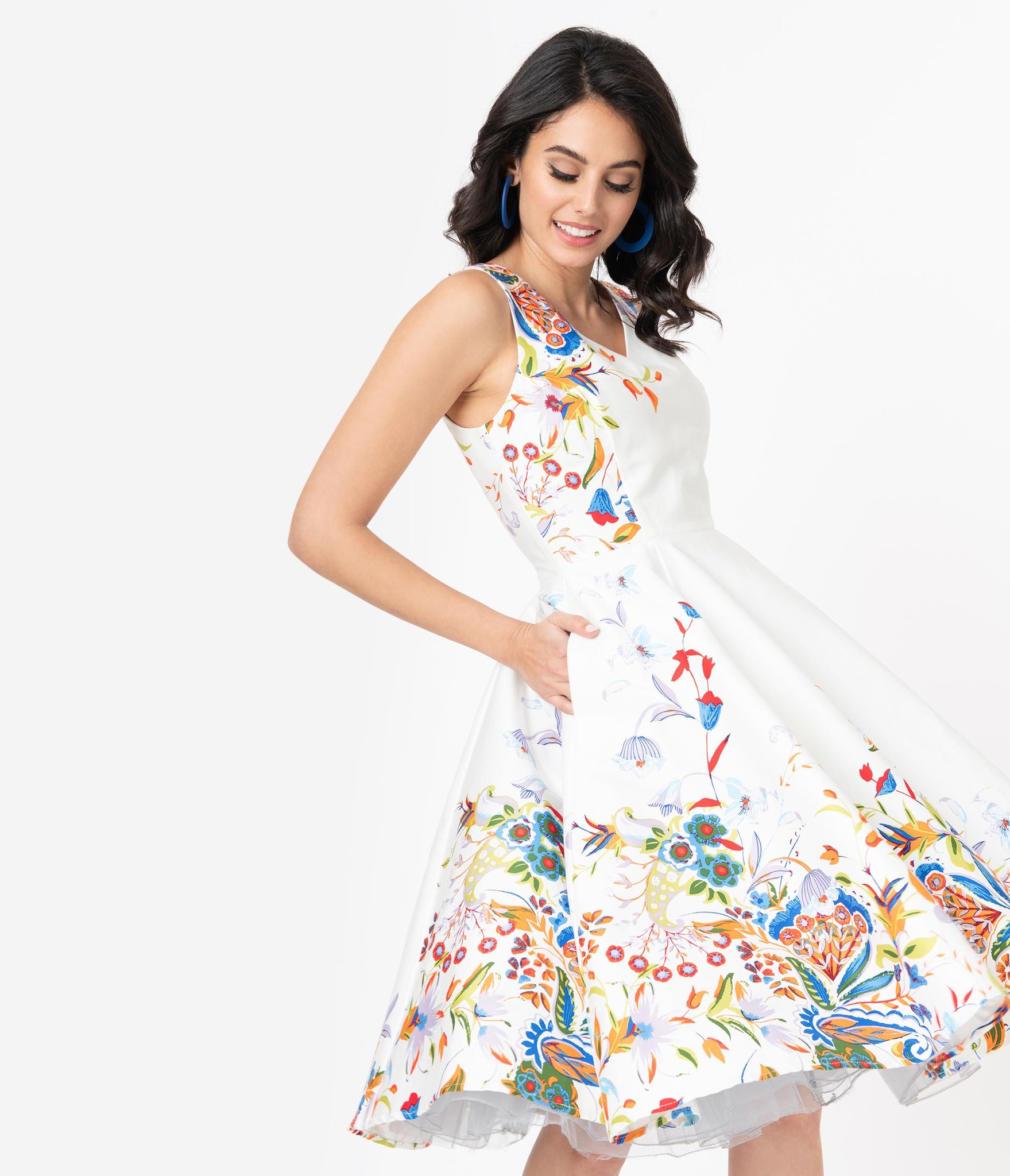 A model wears a knee-length white dress with colorful floral embroidery all around the skirt and up the bodice. The dress is from Modcloth alternative clothing brand Unique Vintage from Europe.