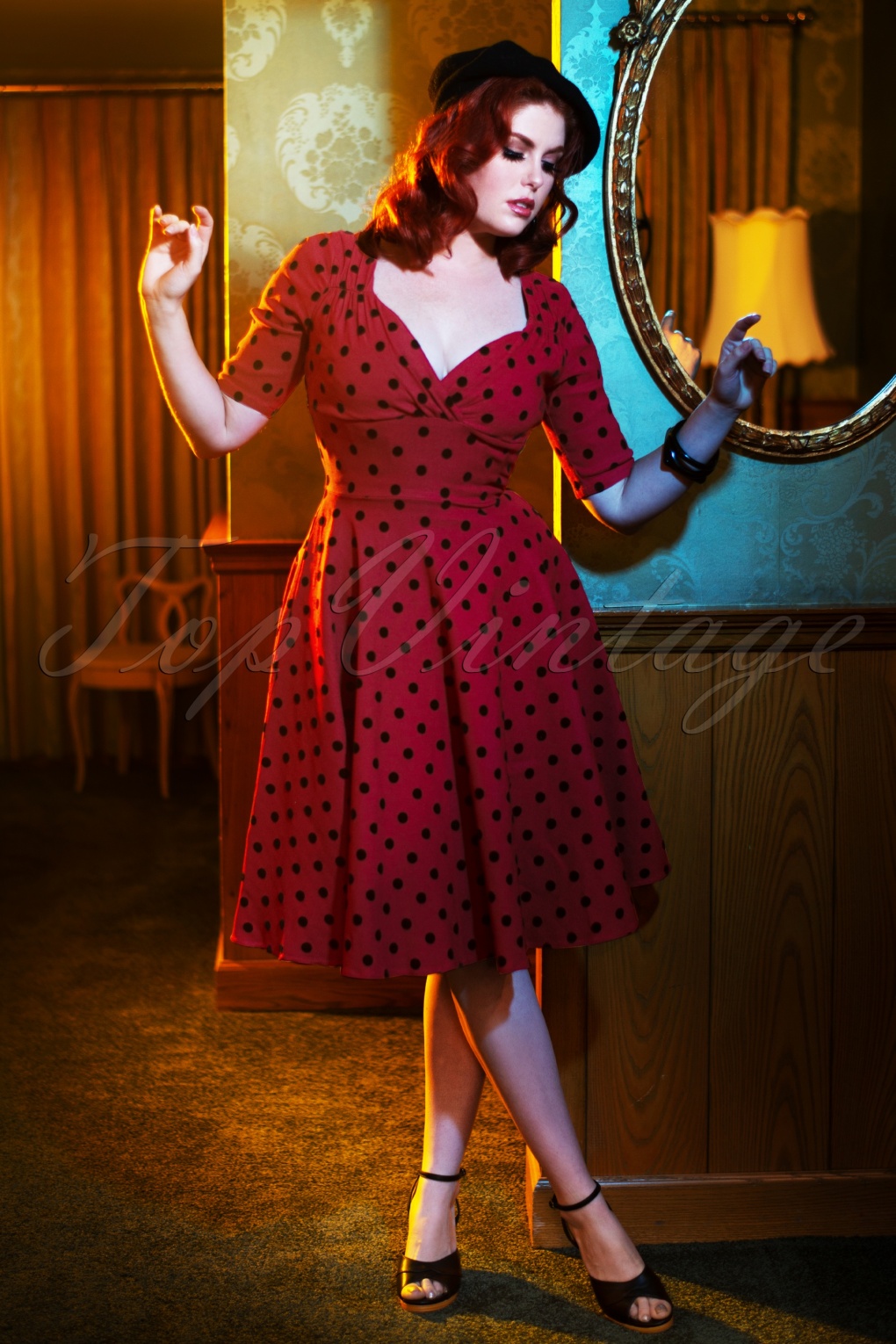 A model wears a vintage a red and black polka dot vintage style dress. She's posed in a dark, warmly lit-room in front of a mirror and walls covered in patterned wallpaper.
