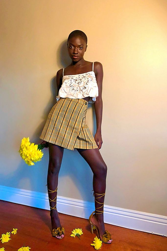 UO Patterned Pleated Mini Skirt paired with a cropped white tank top and strappy sandals. The model is also holding a bouquet of yellow flowers