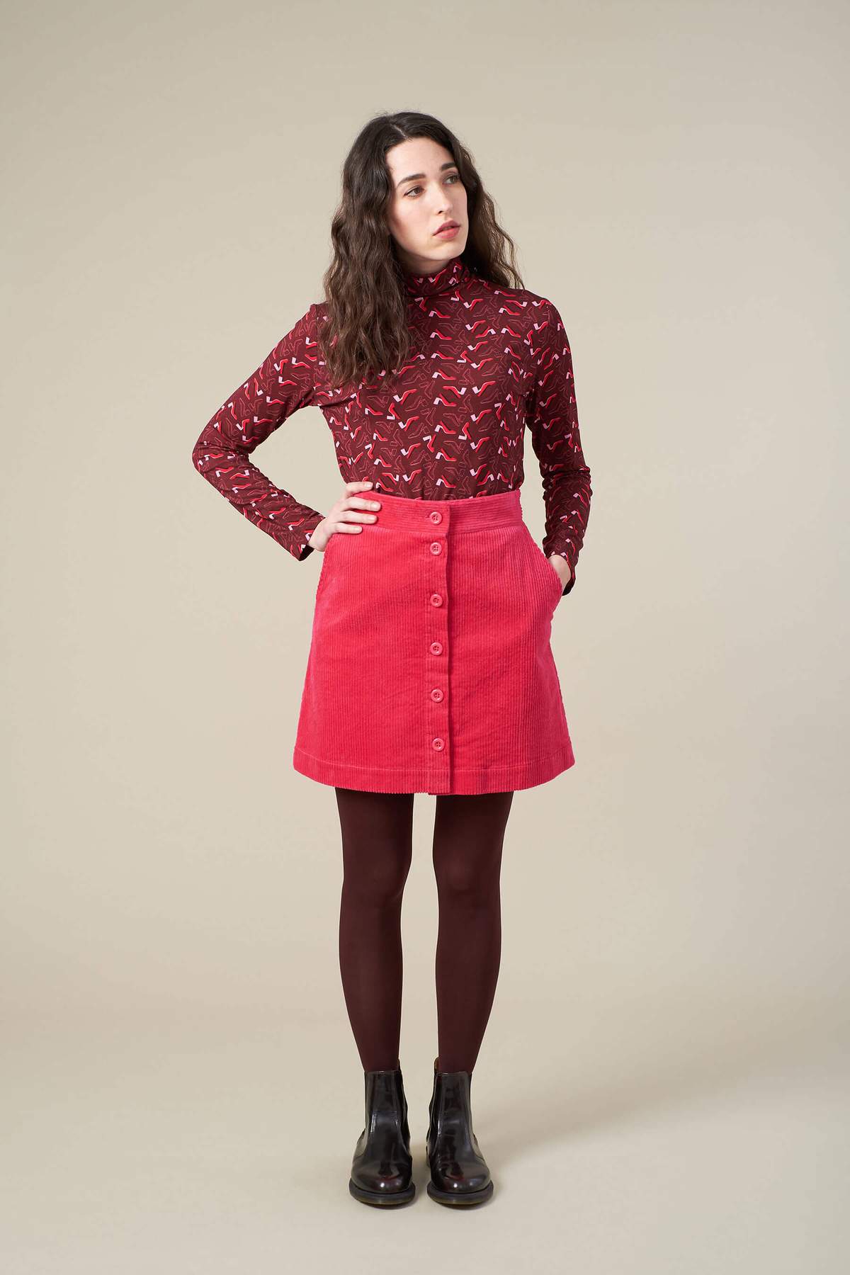 A model wears a pink corduroy skirt paired with a darker maroon red long-sleeve patterned top and black tights. She stands with her hand on her hip, looking away from the camera.