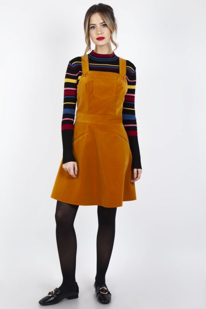 A model wears a gold colored velvet Corduroy Overall dress over a long-sleeved stripped shirt and black leggings. The outfit is from European Modcloth alternative clothing brand Voodoo Vixen.