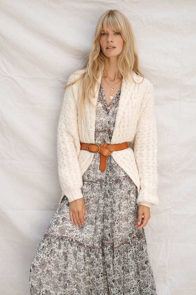 A woman with blonde hair wears a white cardigan secured with a leather belt over a Tiered Maxi Dress from Anthropologie 