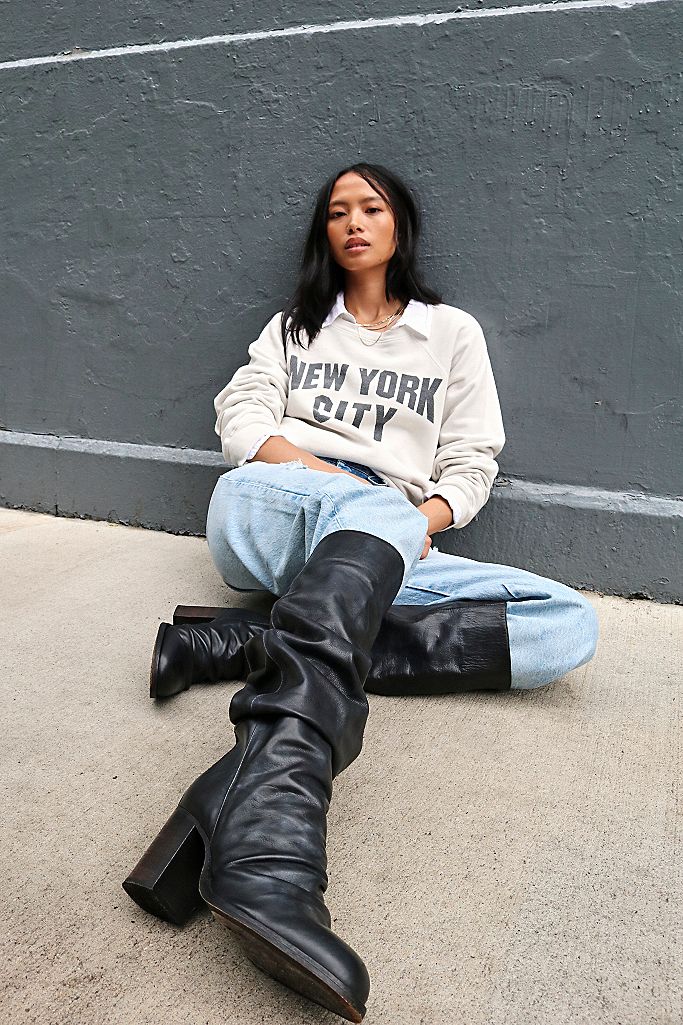 A young woman wearing a white sweatshirt that says New York City in black lettering, and jeans tucked into Elle Tall Slouch Boots. She is sitting on the street leaning against a wall.