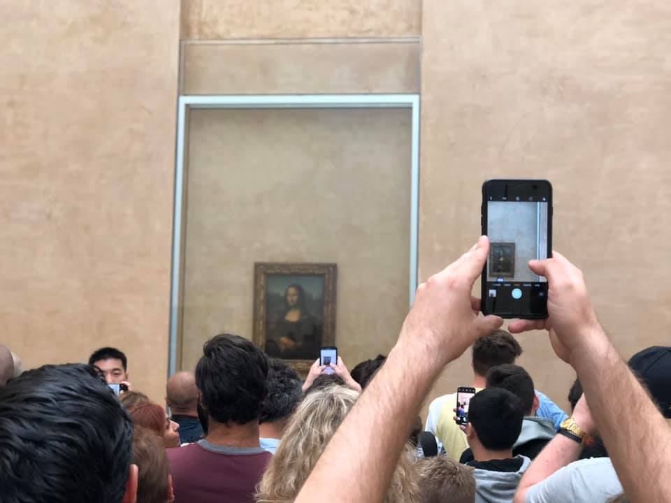 A picture of the Mona Lisa painting in the background, obscured by thick glass and dozens of the back of visitors' heads. One man holds up an iPhone, taking a picture of the painting.