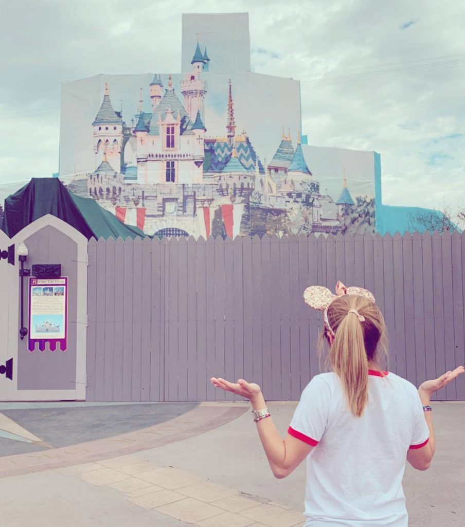 A girl stands with her back tot he camera, looking at the "Cinderella castle", which is under construction and obscured completely by scaffolding that's covered with a large picture of the exterior of the castle.