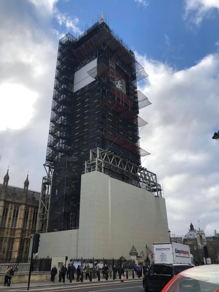 The Big Ben clocktower covered completely by construction scaffolding, standing tall against a blue cloudy sky.