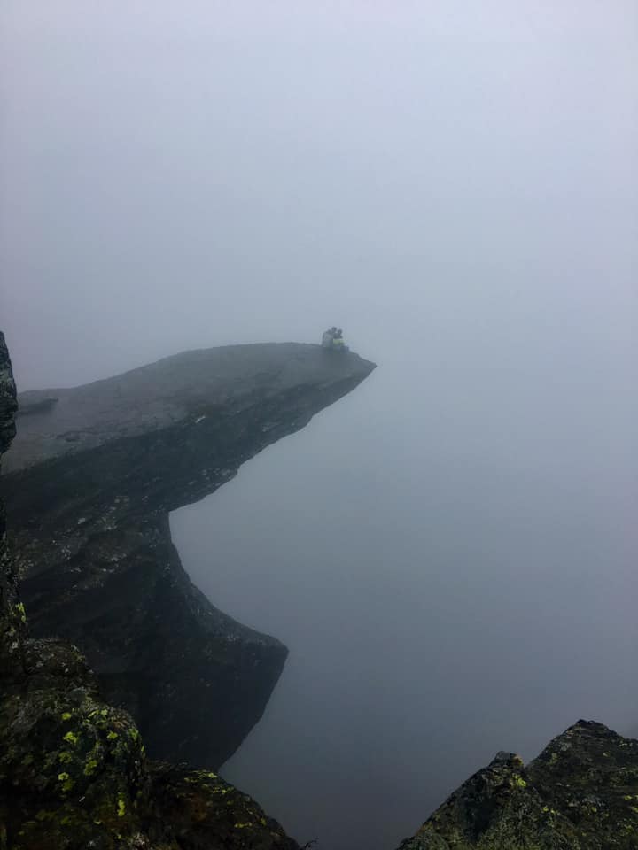 A couple sits at the edge of a cliff overlooking Trolltunga Valley, but the gorgeous view is completely obscured by dense fog.