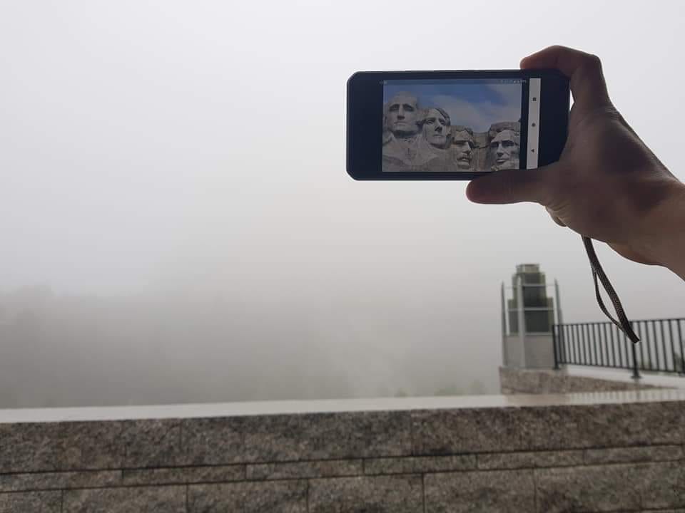 A person holds an iPhone in front of the camera, showing a picture of Mount Rushmore. The background, where Mount Rushmore should be, is obscured by thick fog.