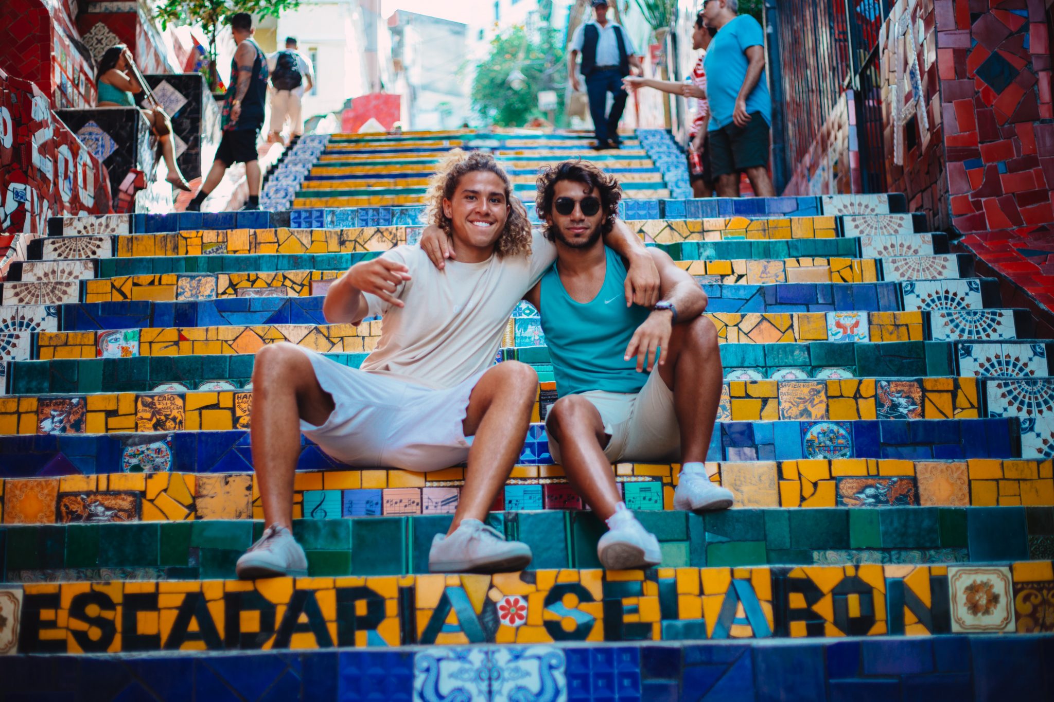 Two men pose with their arms around each other, sitting on the Escadaria Selarón staircase in Rio de Janeiro. The stairs are mostly empty except for a few other tourists towards the top.