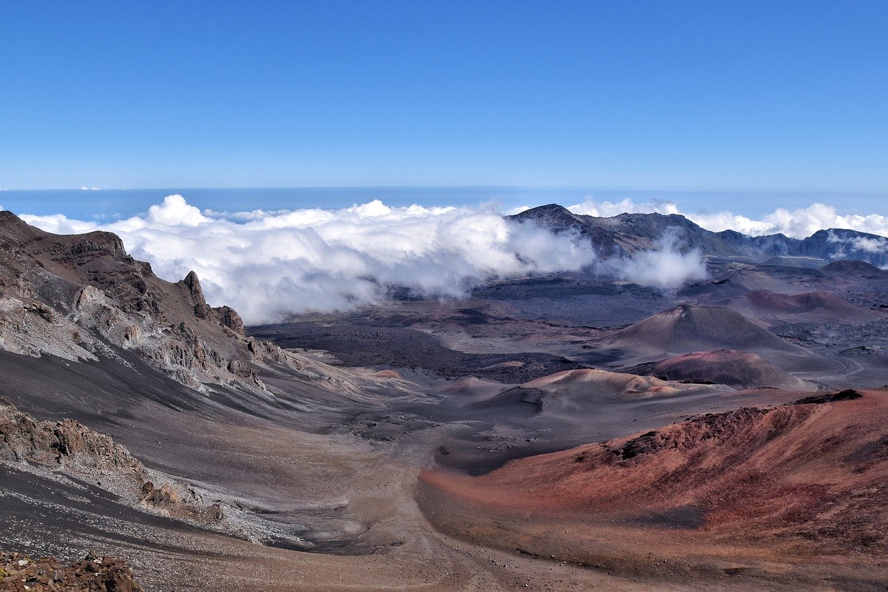 A professional photo of the Haleakala Volcano in Maui on a clear day. The sky is clear blue with low clouds over the mountaintops.