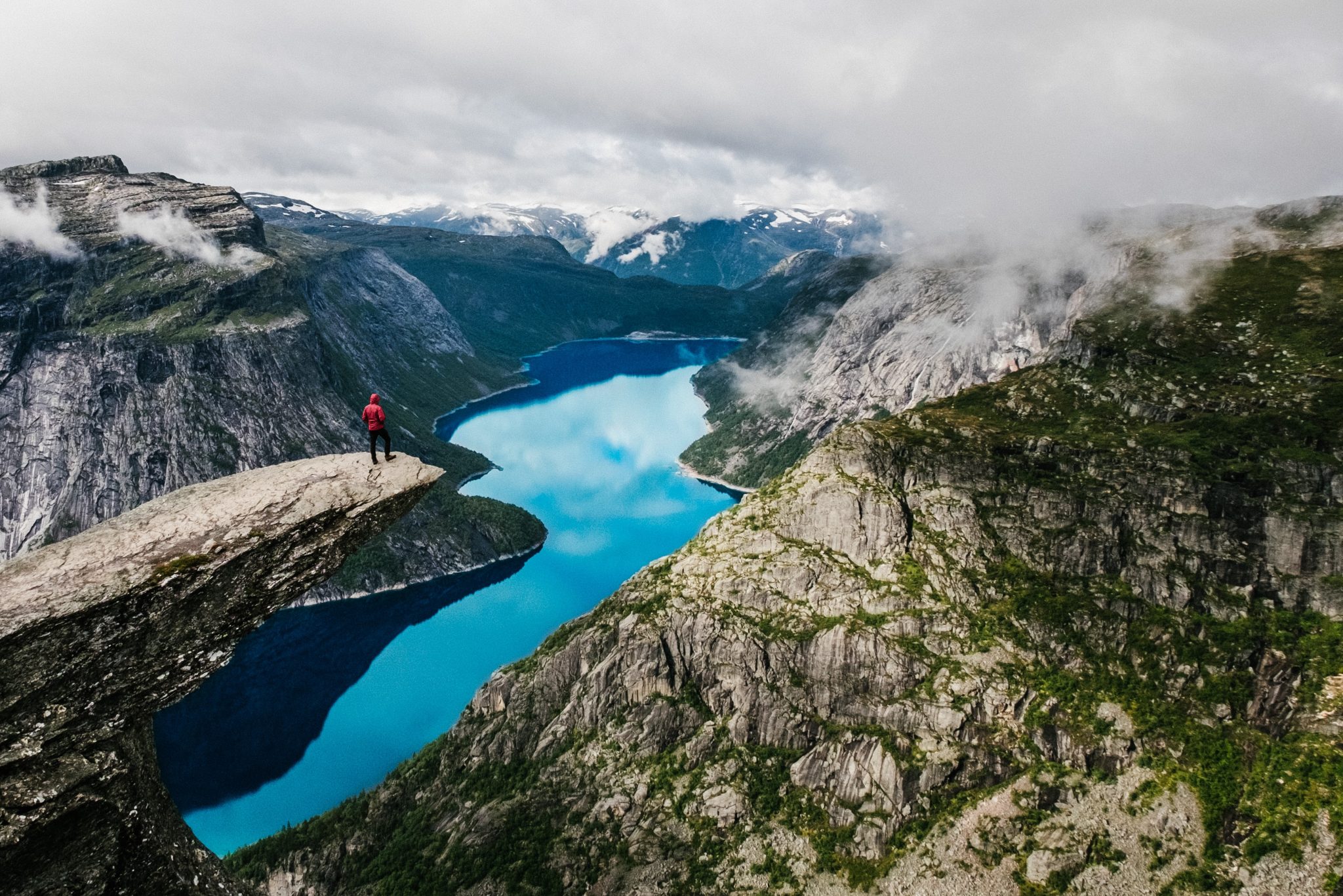 A professional photo of the Trolltunga passage in Norway. A man stands at the edge of a giant cliff overlooking and mountainous valley with a crystal clear blue river snaking through.