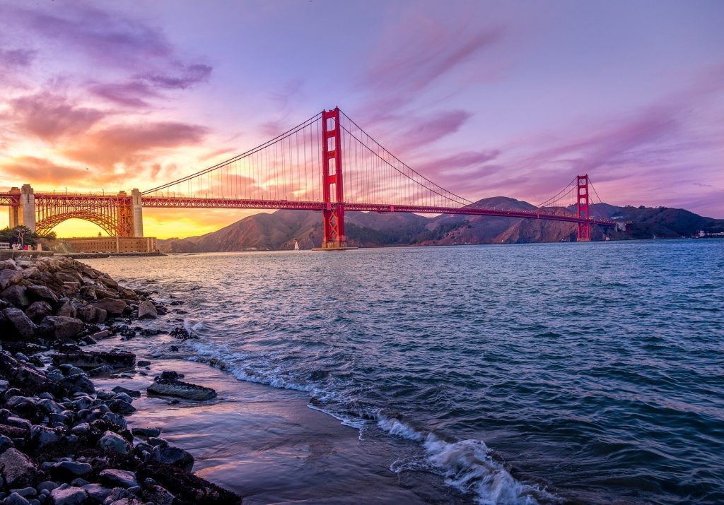 A professional photograph of the Golden Gate Bridge in San Francisco Bay at sunset. The sky is changing colors from rich golds and orange to pinks, purples, and blues.