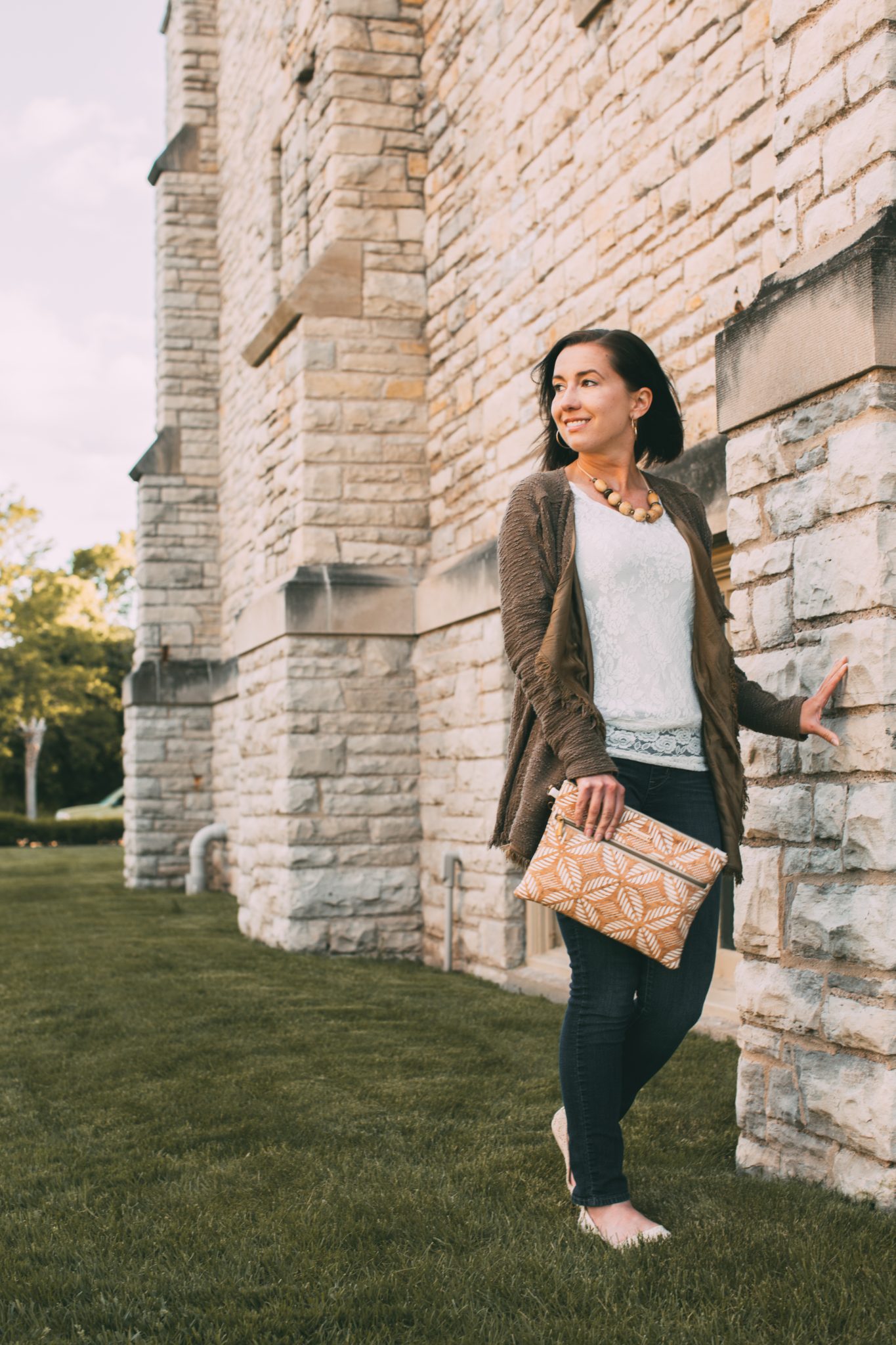 A woman poses outdoors next to a stone wall. She's wearing jeans, a white shirt, and brown cardigan sweater. She's holding the Carry Courage tablet clutch at her side.