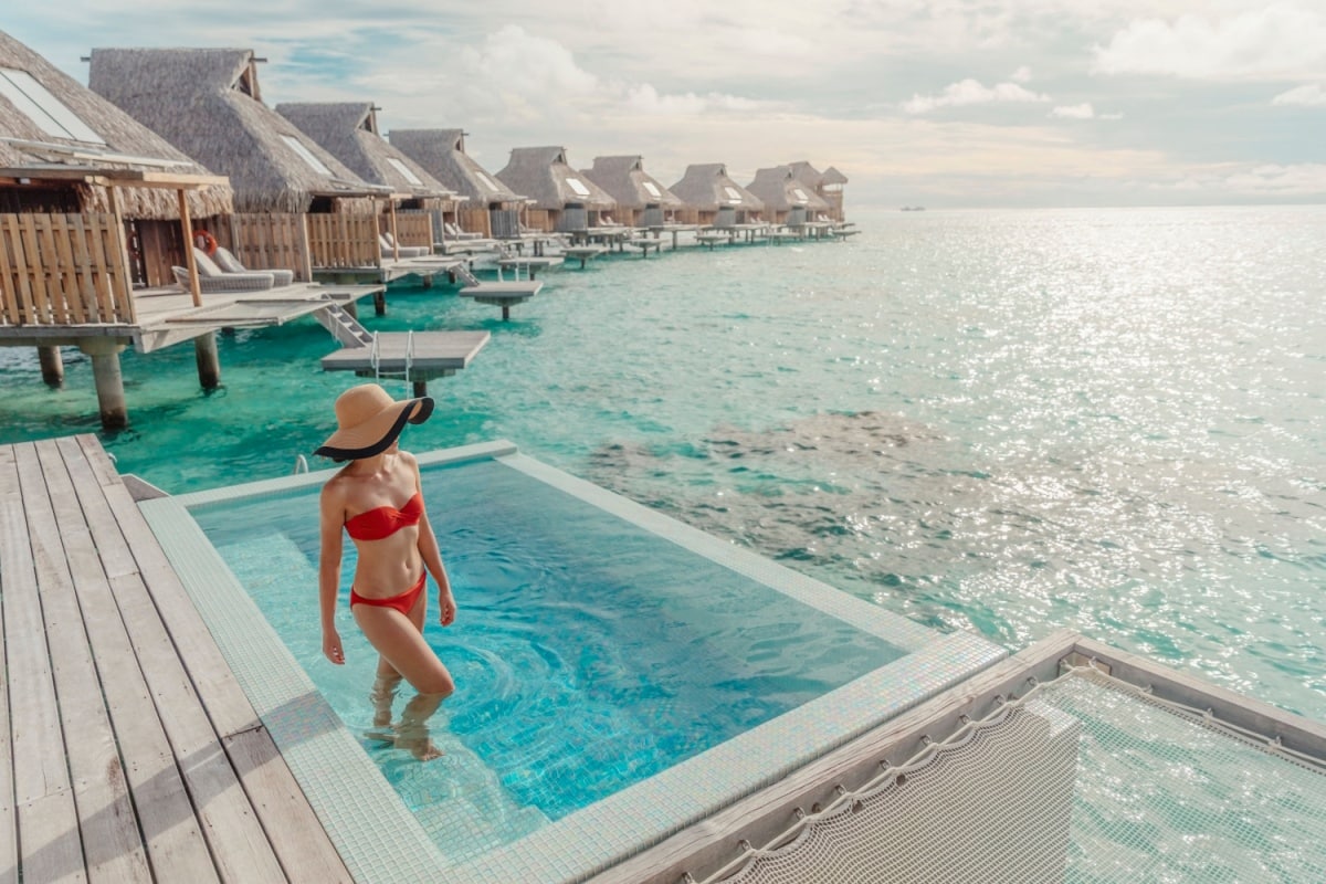 Luxury hotel woman relaxing in infinity swimming pool of private overwater bungalow suite enjoying summer vacation. Tourist with hat and red swimsuit in water sunbathing.