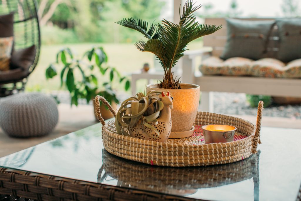 A decorative ratan tray is placed on the glass top of an outdoor table. The tray is filled with two planters and a lit citronella candle in a small metal container.