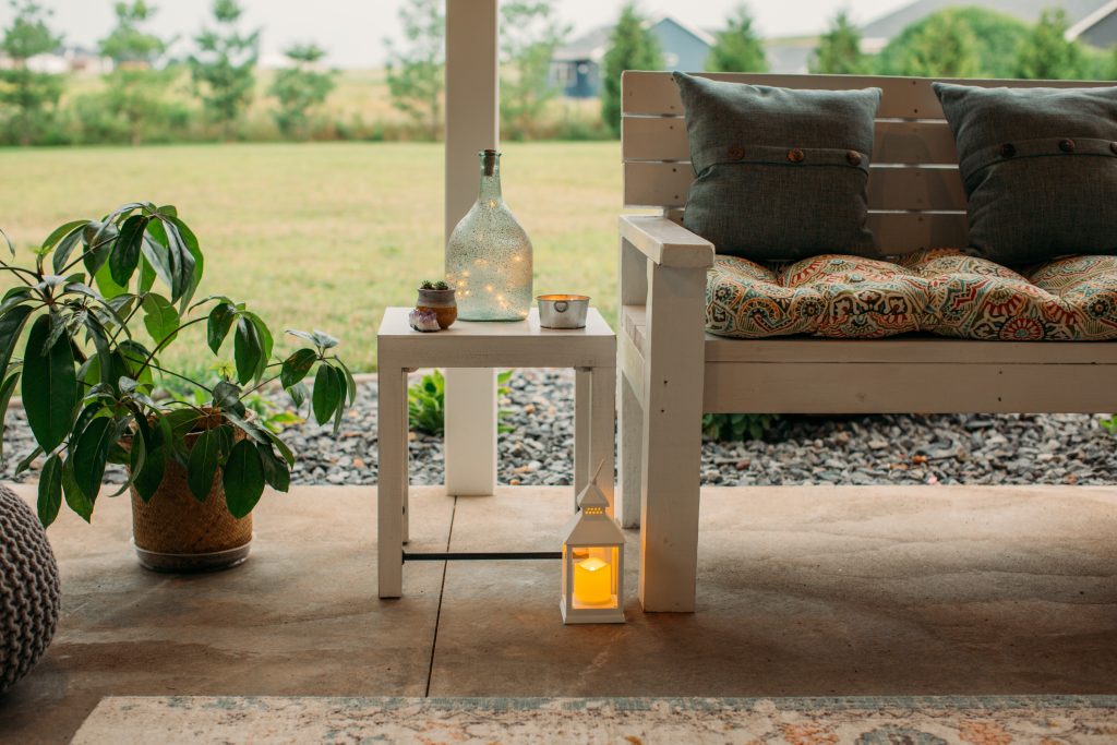 Small decor details around the backyard living space -- a white wooden bench with pillows, small side table, and small candle lanterns filled with flameless candles.