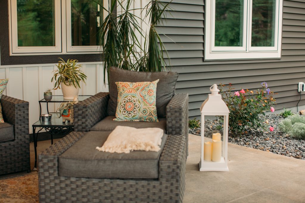 A white decorative lantern sits on the ground next to an outdoor chair. It's filled with three candles that glow a warm, soft light.