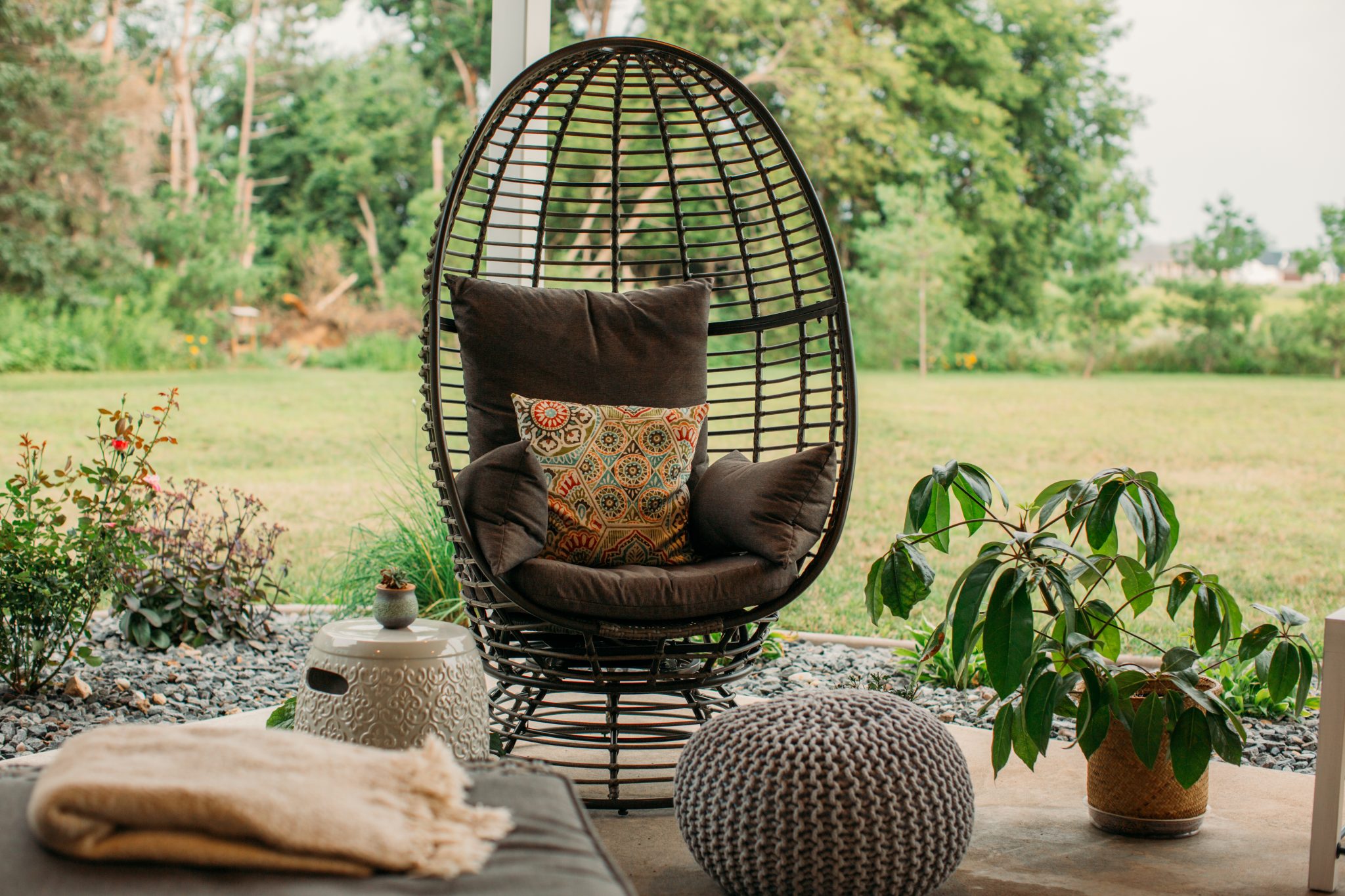 A wicker egg chair with cozy seat cushions and pillows, outside on a cozy backyard patio space.