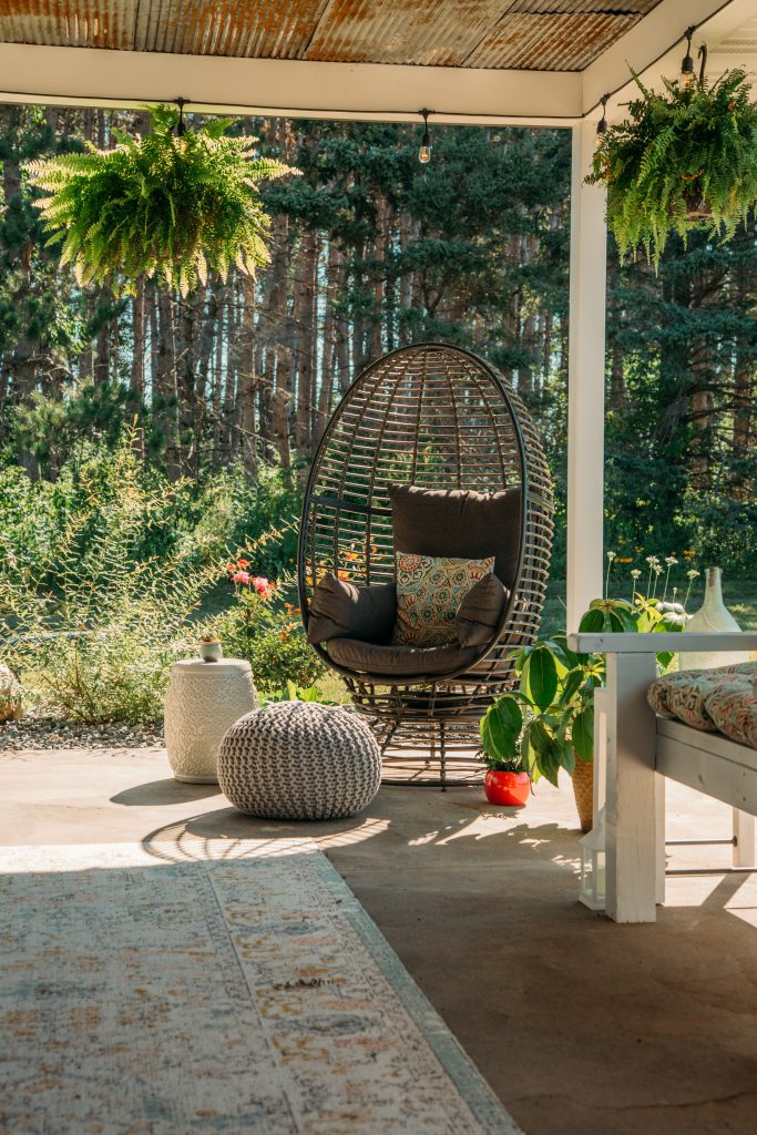 An wicker egg chair sits on the corner of an outdoor patio, surrounded by planters and an ottoman pouf.