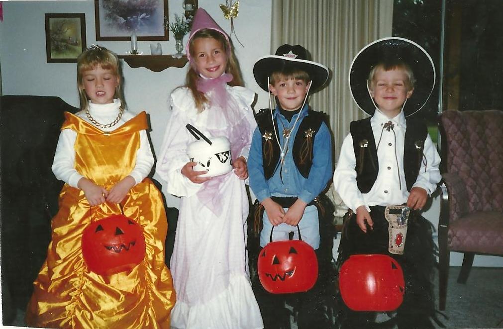 An older film photo of four children dressed up for Halloween. Two little girls on the left are dressed up as princesses, and two little boys on the right are dressed up as cowboys.