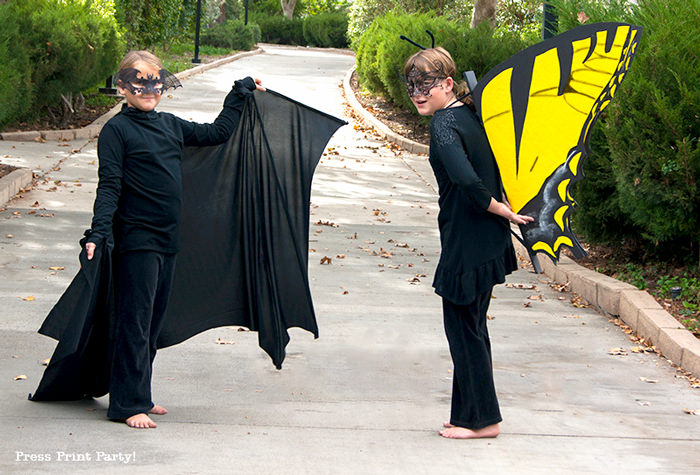Two kids pose on a sidewalk, dressed in all black as a bat and butterfly. The butterfly costume has bright yellow wing.