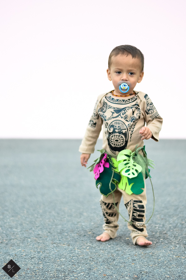 A young boy is dressed as Maui from Moana -- he wears a full-body tan onesie with a tattooed pattern all over, and a tropical grass skirt.