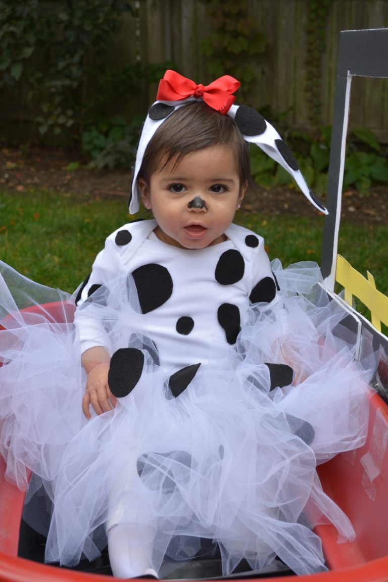 A toddler sits in a wagon dressed as a cute little cow for Halloween. She's wearing a black-spotted onesie, white tulle skirt, and a cow ears headband with a white bow.