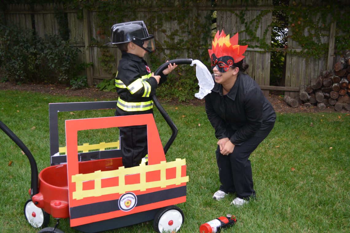 A young child dressed as a firefighter plays in the back of a wagon, spraying a woman dressed as fire with a "firehose".