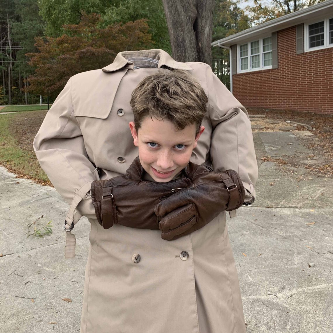 A young boy is dressed in a brilliant DIY Halloween costume. He looks headless, wearing a long beige trench coat and brown cloves, his head floating in the middle.