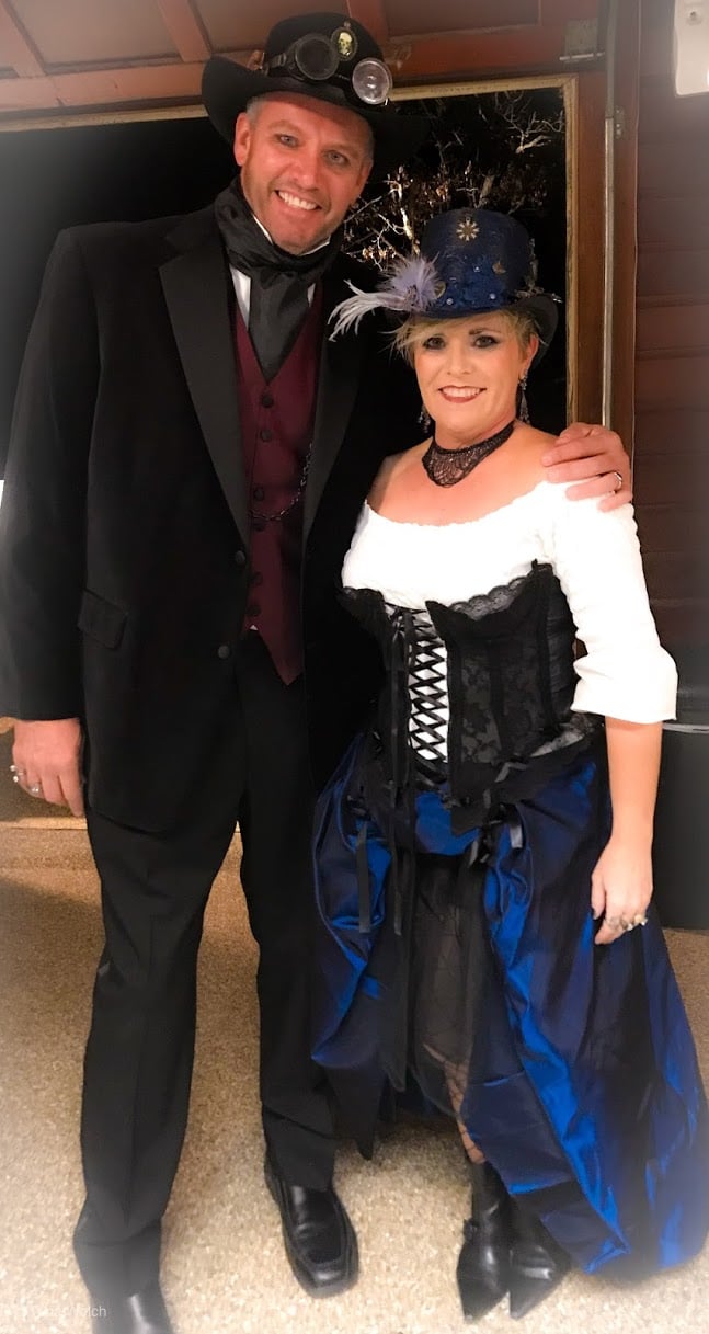 A couple dressed in steampunk-themed Halloween costumes. The man wears a black velvet suit, red vest, and top had. The woman wears a black corset over a white off-the-shoulder blouse and blue satin skirt.