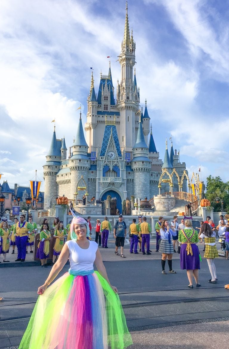A woman poses in front of Cinderella's castle in Disney, and she's wearing a DIY unicorn costume made from a rainbow skirt, white top, pastel colored wig, and a unicorn horn headband.
