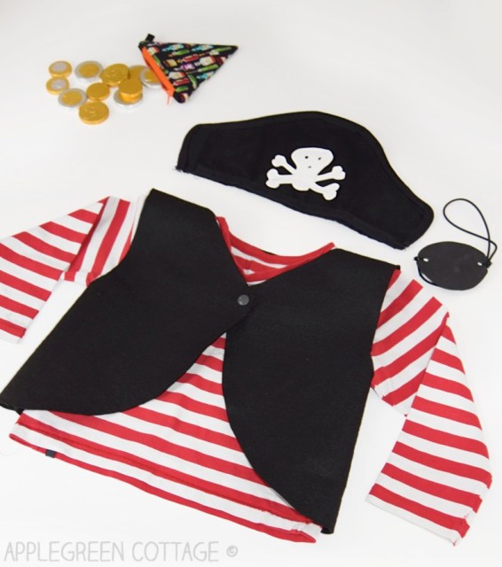 A child's pirate costume - red and white striped shirt, black vest, pirate's hat, and eye patch - all laid out on a white surface. A little pouch of "loot" (chocolate gold coins) sits next to the clothing.