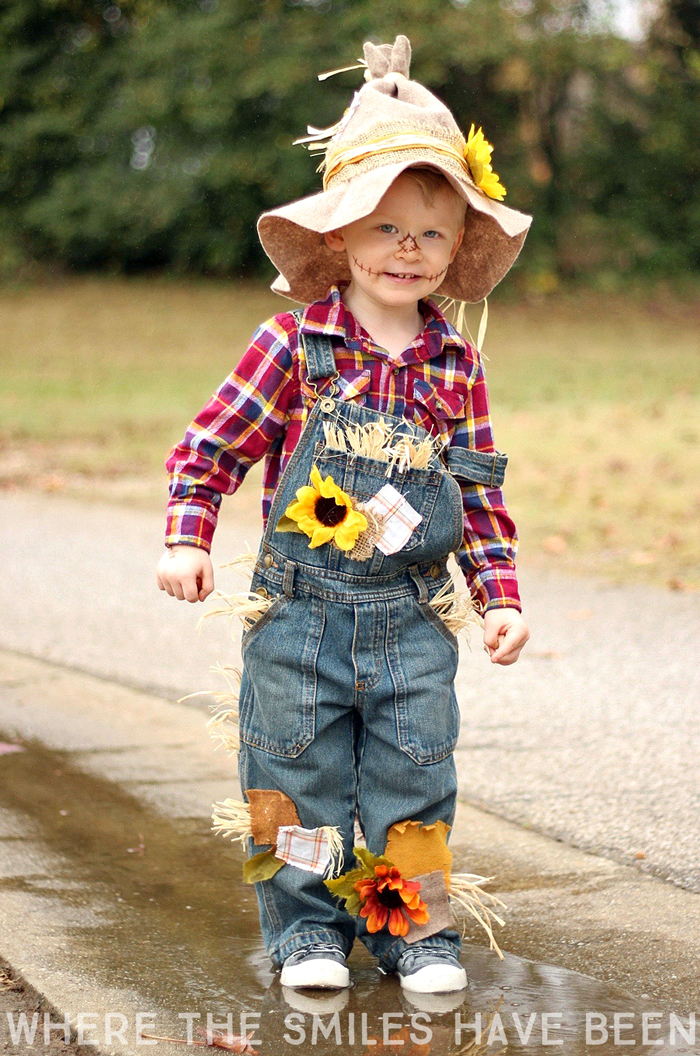 A little boy is dressed in a DIY scarecrow costume, wearing a red plaid shirt, patch-work overalls, and a little straw hat.