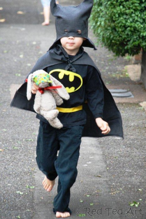 A cute little boy walks down the stressed dressed as Batman, holding a stuffed bunny toy.