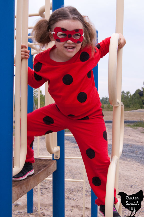 A little girl plays on a jungle gym, dressed as a ladybug. She's wearing a red sweatshirt and pants dotted with black dots, and a DIY red and black mask over her eyes.