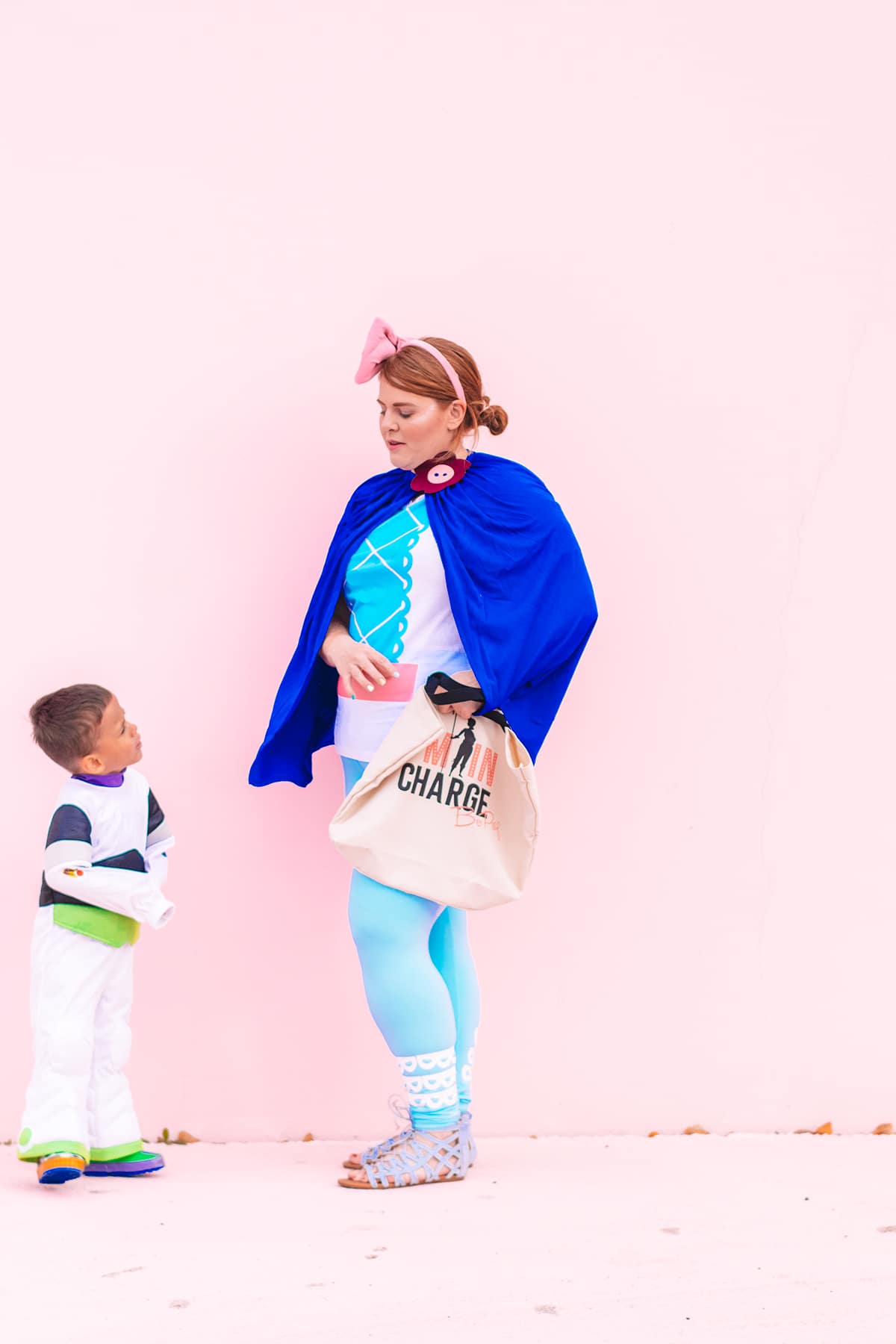 A woman is dressed a Bo Peep, wearing a DIY'd shirt created with a Cricut. She poses with a little boy dressed as Buzz Lightyear in front of a pastel pink wall.