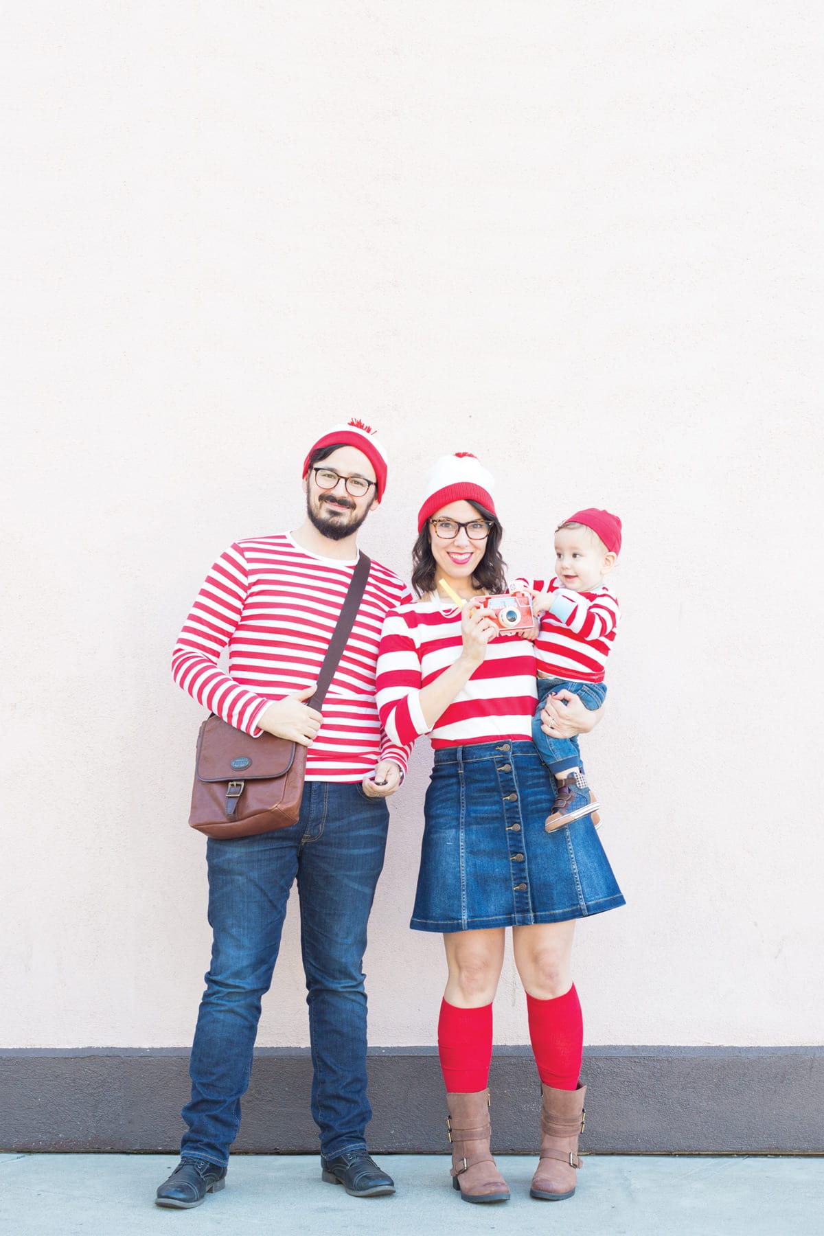 A family of three - mom, dad, and baby - dressed as Where's Waldo in red and white striped shirts, jeans, and red and white hats.
