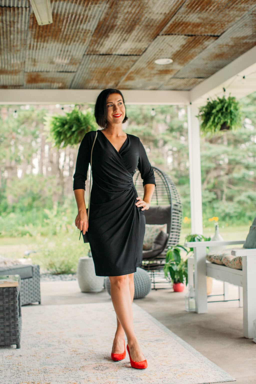 Lindsey wearing a black Anatomie wrap dress with red suede pumps and black wallet on a chain