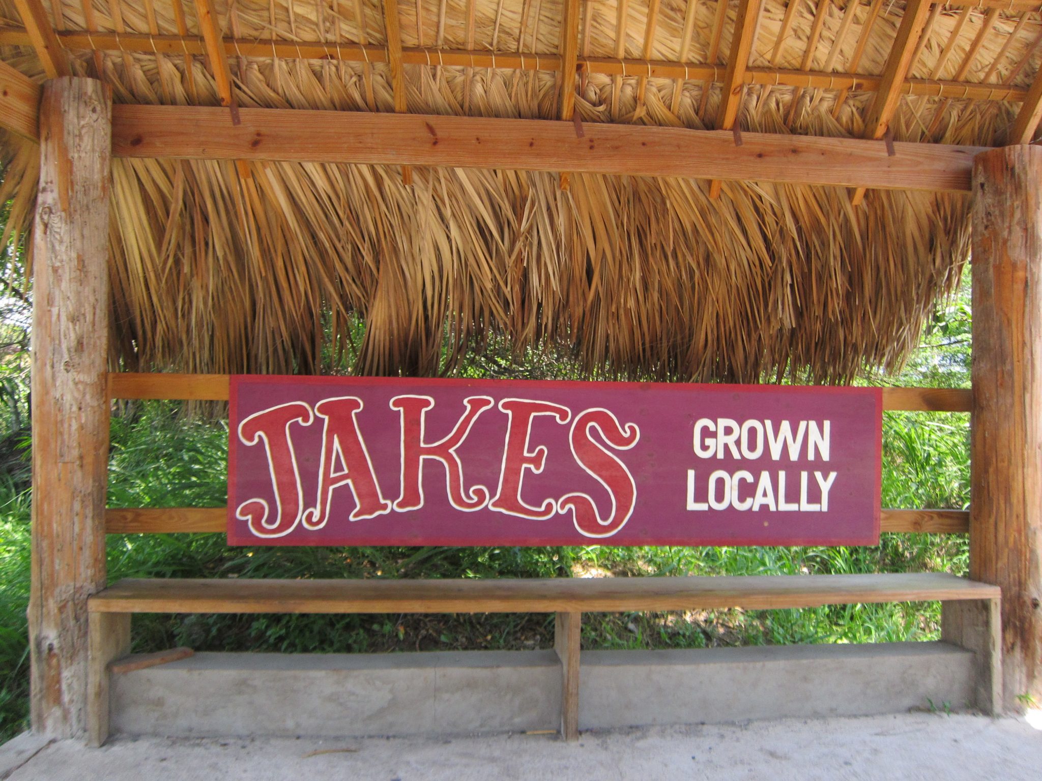 A shaded palm-branch covered bench displays a sign that says "Jakes Grown Locally" in Jamaica.