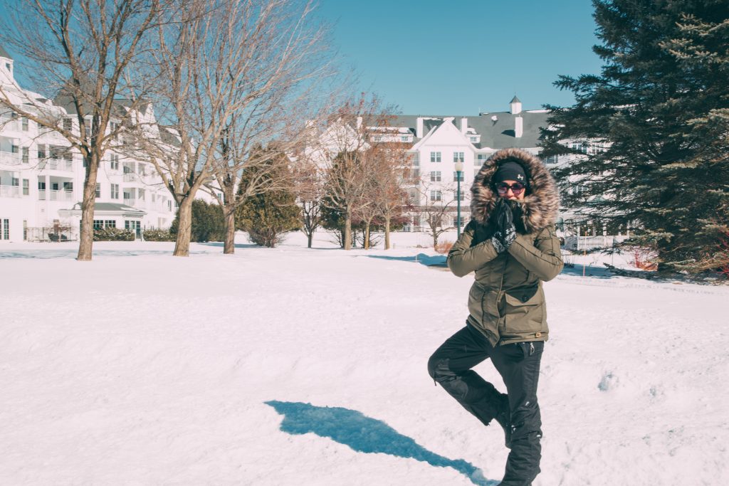 A woman dressed in a heavy winter coat and snow pants stands in a yoga pose in the middle of a snow-covered lawn. Buildings of the Osthoff Resort in Elkhart Lake, WI stand behind bare trees in the background.