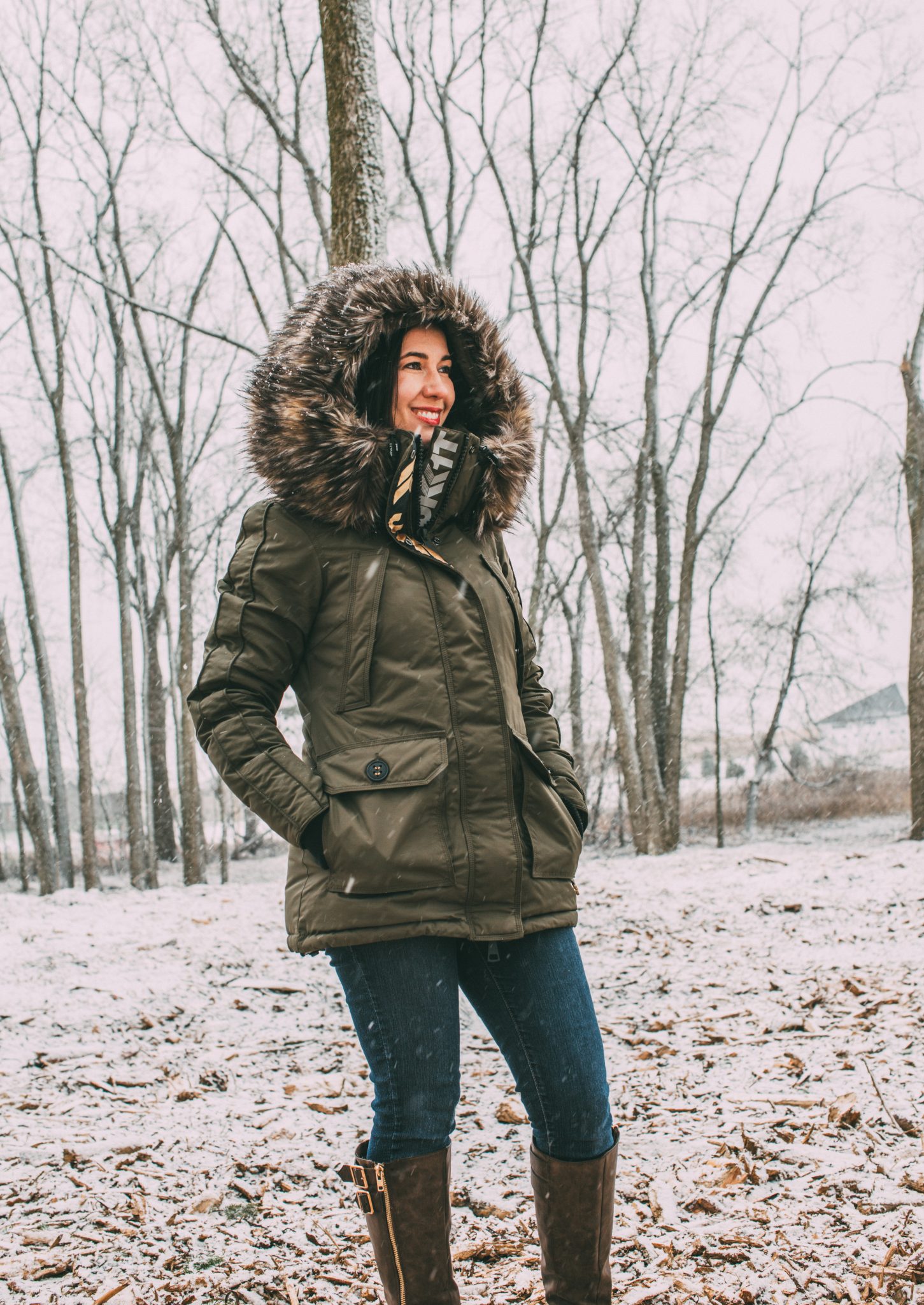 A woman stands in a snowy field wearing a cozy olive green winter jacket with a thick faux fur hood.