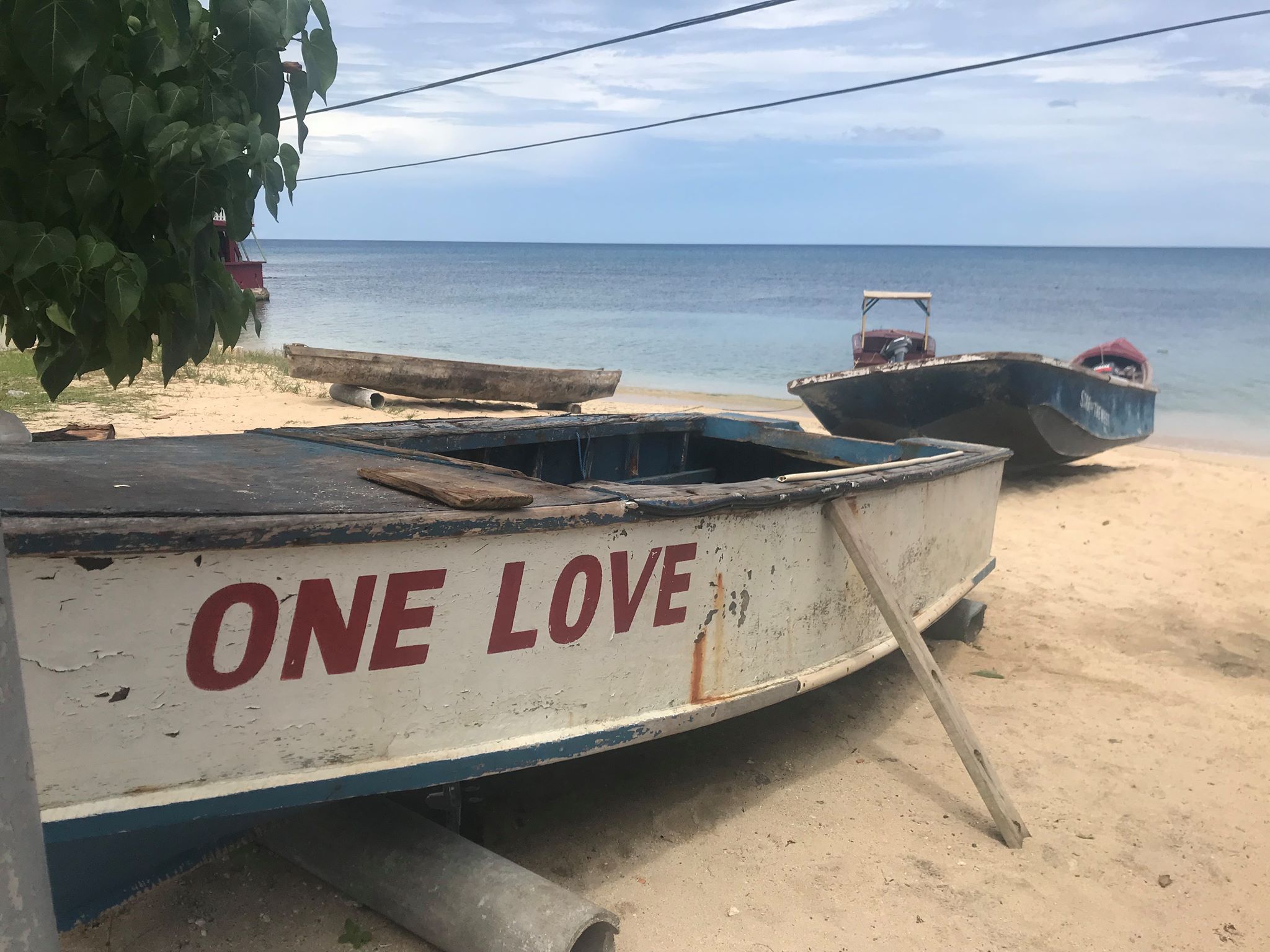 Old wooden boats rest on the sandy beach outside Judy's House in Negril, Jamaica. The boat in the foreground is painted with chopped white and blue paint, and says "one love" in red letters on the side.